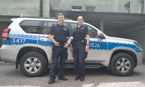 dwoje umundurowanych policjantów stoi przy oznakowanym radiowozie