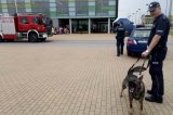 na pierwszym planie widać policjanta przewodnika psa służbowego z psem na smyczy w tle widać wóz strażacki obok policjanta przy radiowozie i z tyłu grupę młodzieży na tle budynku