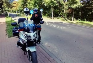 Policjant wydziału Ruchu Drogowego stający przy służbowym motocyklu z laserowym miernikiem prędkości.