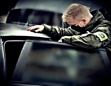 Policjant w cywilnym ubraniu z opaską na ramieniu. Na opasce napis POLICJA. Mężczyzna zagląda do wnętrza samochodu przez otwarte tylne drzwi.