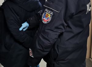 Ręka Policjanta w kurtce policyjnej z emblematem Komendy Miejskiej Policji w Łomży, Policjant trzyma zatrzymanego, który ma założone kajdanki na ręce trzymane z tyłu.