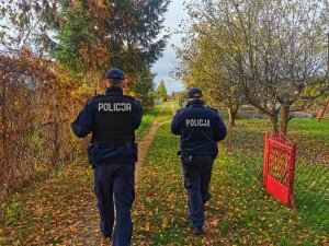 Dwóch  umundurowanych policjantów idzie po ścieżce na ogródkach działkowych