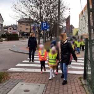 Grupa dzieci, idą w parach, mają na sobie kamizelki odblaskowe. Przechodzą przez przejście dla pieszych w obecności policjantki i nauczycielek.