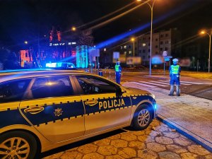 Dwóch umundurowanych policjantów stoi przy przejściu dla pieszych. Za nimi oznakowany radiowóz z włączonymi sygnałami świetlnymi. Pora nocna.