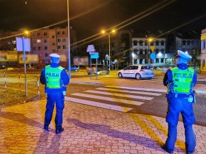 Dwóch umundurowanych policjantów stoi przy przejściu dla pieszych. Pora nocna.