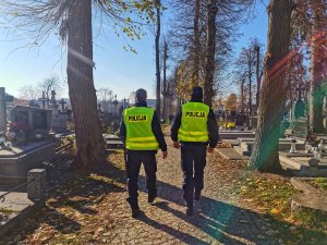 Dwóch umundurowanych policjantów w kamizelkach odblaskowych z napisem policja patrolują cmentarz.