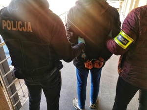 Policjanci w ubraniach cywilnych, jeden z nich ma założoną kamizelkę z napisem policja, a drugi opaskę na ręku z napisem policja. Trzymają zatrzymanego mężczyznę, który ma założone kajdanki na ręce trzymane z tyłu.