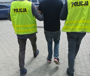 Dwóch policjantów w ubraniach cywilnych, w kamizelkach z napisem policja prowadzą zatrzymanego mężczyznę przez dziedziniec komendy.