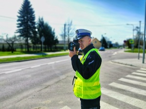 Umundurowany policjant stoi przy jezdni i mierzy prędkość pojazdom.