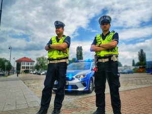 Dwóch umundurowanych policjantów stoi przy przejściu dla pieszych. W tle radiowóz policyjny.