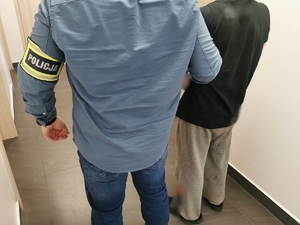 Policjant w ubraniu cywilnym z opaską z napisem Policja założoną na ręku trzyma zatrzymanego mężczyznę, który ma założone kajdanki na ręce trzymane z przodu.
