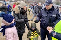Funkcjonariusz Policji przekazuje kobiecie opaskę odblaskową. Poniżej stoi dziecko, które z radością ogląda otrzymany wcześniej od policjanta odblask.