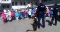 na pierwszym planie dwóch funkcjonariuszy i pies służbowy, policjanci zwróceni do grupy dzieci