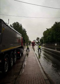 Na zdjęciu widać mokrą nawierzchnię drogi , dwa pojazdy ciężarowe (jeden na parkingu, drugi zjeżdżający na zatoczkę) i dwóch funkcjonariuszy kierujących ruchem