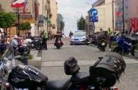 na pierwszym planie górna część motocykla w tle radiowóz policyjny oznakowany pomiędzy kilka innych motocykli i ludzi przechodzących chodnikiem