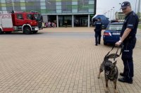 na pierwszym planie widać policjanta przewodnika psa służbowego z psem na smyczy w tle widać wóz strażacki obok policjanta przy radiowozie i z tyłu grupę młodzieży na tle budynku