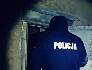 Pora nocna, Policjant tyłem zwrócony do aparatu, policjant ubrany w ciepłą kurtkę z kapturem i napisem POLICJA na plecach, wchodzi do opuszczonego budynku