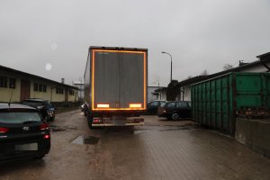 Tył naczepy samochodu ciężarowego na drodze gruntowej. W koło zaparkowane samochody osobowe. Pogoda deszczowa.