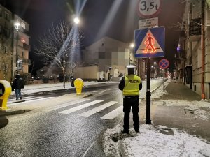 Policjant stojący przy przejściu dla pieszych i znaku drogowym, na pasach mężczyzna , w tle ulica miasta oświetlona latarniami.