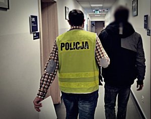 Policjant prowadzący zatrzymanego. Idą przez hol w budynku komendy. Policjant po cywilnemu w żółtej kamizelce z napisem POLICJA