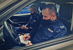 Policjanci Wydziału Ruchu Drogowego siedzący w radiowozie. Piszą w notatnikach służbowych.