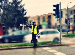Policjant stojący na pasu zieleni, lizakiem służbowym sygnalizuje do pojazdom zjazd z drogi