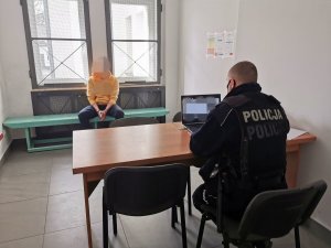 Umundurowany policjant siedzi przy stoliku i pisze na komputerze służbowym. Zatrzymany mężczyzna siedzi na ławce, przodem skierowany do funkcjonariusza.