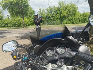 Widoczny służbowy motocykl i stojący przed nim umundurowany policjant, który mierzy prędkość ręcznym miernikiem prędkości.