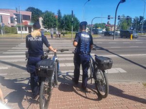 Dwóch umundurowanych policjantów prowadzą rowery służbowe i przechodzą przez przejście dla pieszych.