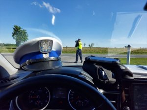 Umundurowany policjant stoi przy jezdni i mierzy prędkość ręcznym miernikiem prędkości. Zdjęcie wykonano z radiowozu. Widoczna czapka policyjna na podszybiu.