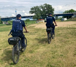Dwóch umundurowanych policjantów jedzie rowerami służbowymi przy plaży w Łomży.