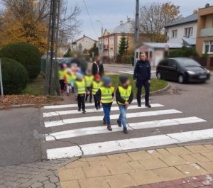 Grupa dzieci, idą w parach, mają na sobie kamizelki odblaskowe. Przechodzą przez przejście dla pieszych w obecności policjantki i nauczycielek.