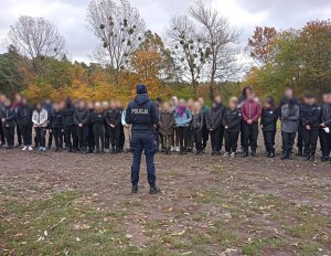 Umundurowana policjantka rozmawia z młodzieżą, która w grupie stoi przed nią. Zdjęcie wykonano w lesie.