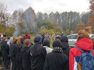 Umundurowana policjantka rozmawia z młodzieżą, która w grupie stoi przed nią. Zdjęcie wykonano w lesie.