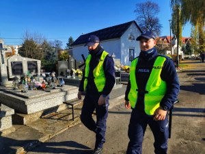 Dwóch umundurowanych policjantów w kamizelkach odblaskowych z napisem policja patrolują cmentarz.