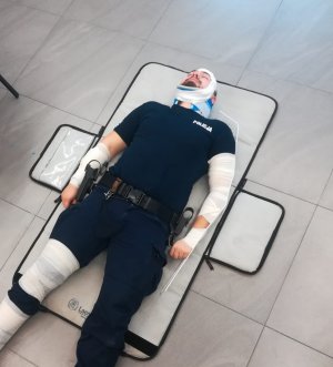 Leżący policjant, który obandażowaną ma nogę i rękę oraz głowę.