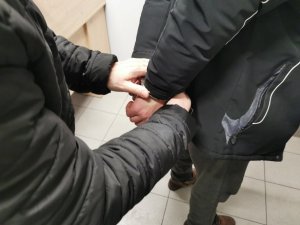 Policjant w ubraniu cywilnym zakłada kajdanki na ręce trzymane z tyłu zatrzymanemu mężczyźnie.