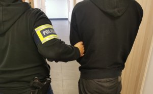 Policjant w ubraniu cywilnym z założoną opaską na ręku z napisem policja trzyma osobę zatrzymaną na korytarzu komendy.