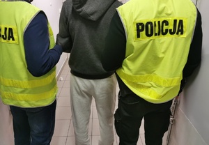 Policjanci w ubraniach cywilnych w kamizelkach z napisem policja trzymają zatrzymanego mężczyznę