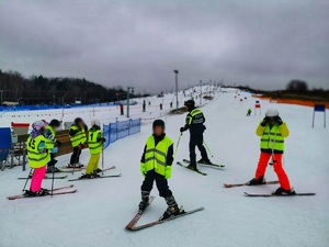 Umundurowany policjant stoi na stoku na nartach. Wokół niego dzieci na nartach.