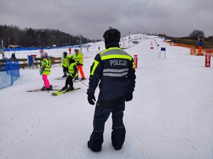 Umundurowany policjant stoi na stoku. Wokół niego dzieci na nartach.