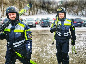 Dwóch umundurowanych policjantów z patrolu narciarskiego idą na stok, niosąc narty. W tle zaparkowane samochody.