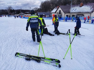 Dwóch umundurowanych policjantów z patrolu narciarskiego udziela pierwszej pomocy przedmedycznej poszkodowanemu narciarzowi, który leży na stoku.