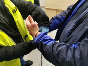 Policjant w ubraniu cywilnym z kamizelką odblaskową z napisem zakłada zespolone kajdanki.zatrzymanemu.