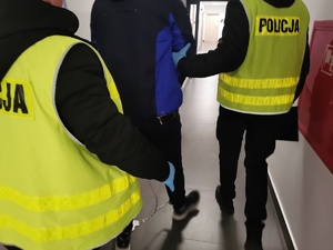 Policjanci w ubraniach cywilnych w kamizelkach odblaskowych z napisem policja prowadzą zatrzymanego w zespolonych kajdankach.