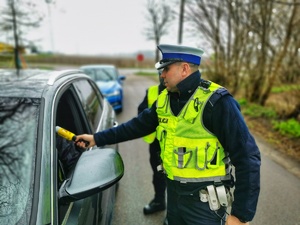 Dwoje umundurowanych policjantów w trakcie kontroli drogowej samochodu osobowego. Policjant przeprowadza badanie stanu trzeźwości kierowcy.