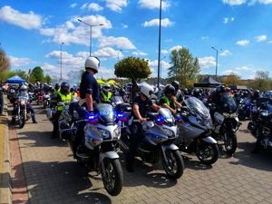 Motocykliści siedzący na motocyklach. Przed nimi policjanci na motocyklach służbowych.