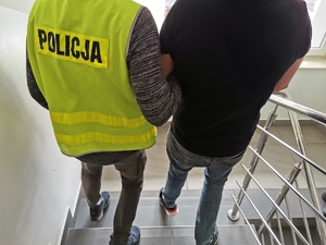 Policjant w ubraniu cywilnym, w kamizelce z napisem policja prowadzi zatrzymanego mężczyznę schodami w komendzie.