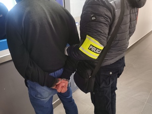 Policjant w ubraniu cywilnym z opaską na ręku z napisem policja trzyma zatrzymanego mężczyznę, który ma założone kajdanki na ręce trzymane z tyłu.