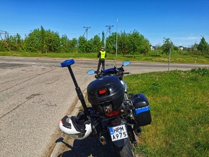 Umundurowany policjant mierzy prędkość pojazdom ręcznym miernikiem prędkości. Widoczny motocykl policyjny.